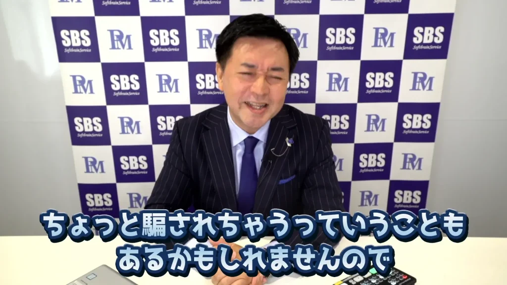 スーツとネクタイ姿の男性が机に座り、笑顔で話している。彼の後ろには「PM」と「SBS」の文字が入ったロゴが描かれた背景がある。画像の下部には日本語の文字が表示されており、彼が研修講師であることを示していると思われる。