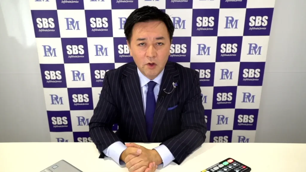 黒いピンストライプのスーツを着た男性が、白い机の前に座り、両手を組んでいる。彼は「SBS」と「RM」のロゴが繰り返し描かれた背景に向かって話している。机の上には、科学的組織営業（科学的組織営業）に欠かせないツールであるリモコンとノートパソコンが置かれている。